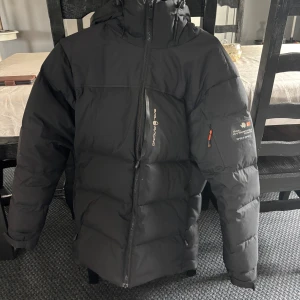 Svart dunjacka Sail Racing herr - Svart dunjacka från Sail Racing med huva och dragkedja. Jackan har logga på bröstet och ärmen, samt en bröstficka med dragkedja. Puffig modell som håller dig varm under kalla dagar. Snygg och sportig look med detaljer i vitt och orange. Inte bästa skicket men funkar absolut för en vinter.