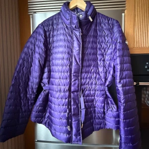 Lila dunjacka från Michael Kors - Snygg lila dunjacka från Michael Kors med quiltad design, hög krage och bälte i midjan. Jackan har dragkedja framtill och långa ärmar. Perfekt för dig som vill ha en lätt och trendig jacka med schysst passform.