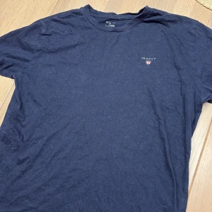Mörkblå t-shirt från GANT - Snygg mörkblå t-shirt från GANT med klassisk rund hals och liten broderad logga på bröstet. Tillverkad i mjuk bomull som känns skön mot huden. Perfekt basic-plagg med stilren vibe och diskret branding.