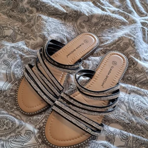 Svarta sandaler med strass från Claudia Ghizzani - Snygga sandaler från Claudia Ghizzani i svart med flera remmar dekorerade med glittrande strass. Platt sula i beige och svart, öppen tå och bekväm slip-in-modell. Perfekta för sommardagar när du vill ha något både stilrent och glammigt.