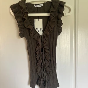 Brun ribbad volangtop från Zara - Säljer en brun ribbad topp från Zara med volanger längs v-ringningen och snörning framtill. Toppen är ärmlös och har en croppad passform som ger en trendig look. Perfekt för dig som gillar detaljer och vill sticka ut lite extra.