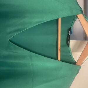 Grön långklänning från ZARA - Snygg grön långklänning från ZARA i ett slätt och glansigt material. Klänningen har v-ringad hals, korta ärmar och en slits på sidorna för extra rörelsefrihet. Perfekt för dig som gillar minimalistisk stil och vill sticka ut med färg. Obsevera. en liten fläck på vänster kläningsarm som möjligtvis går att få bort. Passar stl M-L