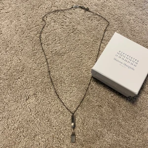 Maison Margiela silver halsband 65cm - Unikt halsband från Maison Margiela i silverfärgad metall med en tvistad, rektangulär berlock som har siffror ingraverade. Kedjan är tunn och har en liten platta med Maison Margiela Paris ingraverat vid låset. Kommer med original ask.