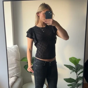 Svart blus med volangärm och mönster - Säljer en svart blus med korta volangärmar och diskret rutmönster. Toppen är croppad och har en feminin siluett med rund halsringning. Materialet känns lätt och har en snygg struktur som gör den extra intressant. Perfekt för dig som gillar stilrena detaljer.