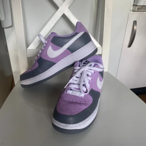 Nike Air Force 1 lila och grå sneakers - Nike Air Force 1 sneakers i en snygg kombination av lila och grått med vita detaljer. Skorna har klassisk låg siluett, vit swoosh och snörning. Ovandelen är i mocka och skinn, med perforerad tå för extra ventilation. Perfekt för dig som vill sticka ut med färg på fötterna. Helt nya!