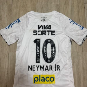Vit Santos Neymar Jr fotbollströja Umbro - Santos FC matchtröja i vitt från Umbro med Neymar Jr och nummer 10 på ryggen. Tröjan har korta ärmar, flera sponsorloggor och klubbmärke på bröstet. Tillverkad i lätt och ventilerande polyester, perfekt för fotbollsplanen eller att bära som supporter.