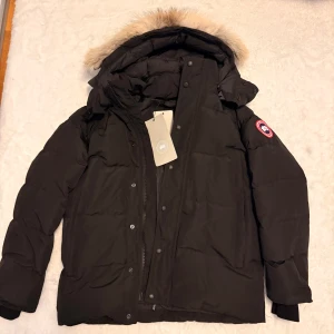 Svart dunjacka från Canada Goose - Säljer en svart dunjacka från Canada Goose med avtagbar huva . Jackan har klassisk patch på ärmen, dragkedja och knappar framtill samt flera fickor. Perfekt för kalla vinterdagar och riktigt snygg streetstil.