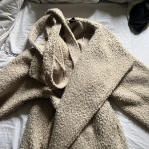 Beige teddyjacka från Gina Tricot - Mysig beige teddyjacka från Gina Tricot i storlek S. Jackan har en oversized passform, bred krage och knytskärp i midjan. Tillverkad i ett fluffigt teddy-material som håller dig varm under kyliga dagar. Perfekt för dig som gillar en trendig och chill look.