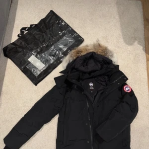 Svart Canada Goose dunjacka med päls - Säljer en svart Canada Goose dunjacka i modell wyndham . Jackan har en stor huva med äkta päls, klassisk röd logotyp på ärmen och dragkedja framtill. Perfekt för kalla vinterdagar och riktigt snygg streetstil.