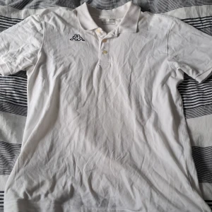 Vit Kappa pikétröja med broderad logga - Vit pikétröja från Kappa i bomull med klassisk krage och knappar framtill. Broderad Kappa-logga på bröstet och tryckt Kappa-text på baksidan av kragen. Kortärmad och stilren design som passar perfekt till jeans eller shorts.
