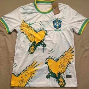 Vit Brasilien fotbollströja Nike - Snygg vit Brasilien fotbollströja från Nike med stora gula fåglar och gröna detaljer. Tröjan har korta ärmar, grönt och gult kantband vid hals och ärmslut samt det klassiska CBF-emblemet broderat på bröstet. Tillverkad i lätt och ventilerande material.