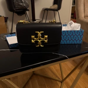 Svart axelväska från Tory Burch - Stilren svart axelväska från Tory Burch i skinn med en chunky guldfärgad kedja och stort guldfärgat logospänne framtill. Väskan har en klassisk, boxig form och levereras i originalkartong. Perfekt för dig som vill ha en snygg och trendig accessoar.