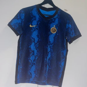 Inter Milan blå matchtröja Nike - Inter Milan fotbollströja från Nike med blått och svart ormmönster, klubbmärke och Nike-logga i guld på bröstet. Tröjan är kortärmad och har en sportig passform med rund hals. Perfekt för dig som vill sticka ut på planen eller på läktaren.