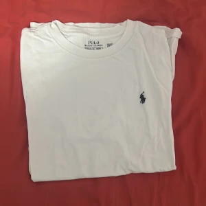 Vit t-shirt från Polo Ralph Lauren - Vit klassisk t-shirt från Polo Ralph Lauren med rund halsringning och ikoniska broderade loggan på bröstet. Tillverkad i mjuk bomull för en skön känsla. Perfekt basic-plagg med stilren design. Storlek 14-16 år