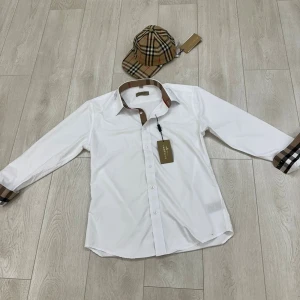 Vit Burberry skjorta med rutig krage - Snygg vit skjorta från Burberry med klassisk rutig detalj på krage och manschetter. Skjortan har lång ärm, knappar framtill och diskret broderad logga på bröstet. Perfekt för dig som gillar stilrena plagg med lyxig touch och ikoniska mönster.