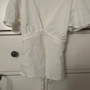 Vit omlottopp från Gina Tricot - Säljer en vit omlottopp från Gina Tricot i storlek XS. Toppen har korta, vida ärmar och knytband i midjan för en snygg passform. Tillverkad i ett mjukt och lätt material som känns skönt mot huden. Perfekt att styla med jeans eller kjol.