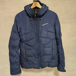 Mörkblå Peak Performance dunjacka - Peak Performance dunjacka i mörkblått med snygg quiltad design och huva med snörning. Jackan har dragkedja framtill, två sidofickor med dragkedja och logga på bröstet. Perfekt för kalla dagar och har en clean, sportig look.