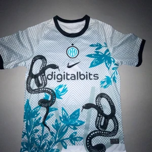 Inter Milan x Nike fotbollströja ormmönster - Inter Milan fotbollströja från Nike med unikt ormmönster och blåa blommor. Tröjan är vit med svarta och blå detaljer, har korta ärmar och svart krage. Digitalbits sponsortryck på bröstet och klubbmärke i blått. Tillverkad i lätt och ventilerande material.