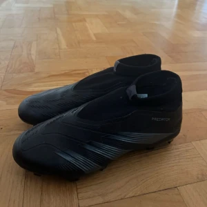 Svarta Adidas Predator fotbollsskor - Säljer ett par svarta Adidas Predator fotbollsskor med strumpliknande skaft och mönstrad ovansida. Skorna har platt sula med dobbar för gräsplan och en snygg, modern design. Perfekt för dig som vill ha grym touch på planen.