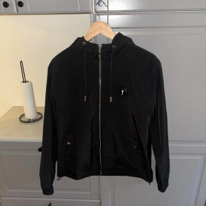 Svart vindjacka från Trapstar - Svart vindjacka från Trapstar med huva, hel dragkedja och snörning i halsen. Jackan har logga på bröstet och stor Trapstar-text på ryggen. Två fickor framtill med knappstängning. Perfekt för dig som gillar streetwear och vill sticka ut.