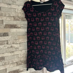 Svart kortklänning med rosa kronor - Svart kortklänning från me&i med rosa kronmönster över hela klänningen. Modellen har rund halsringning och korta ärmar. Tyget är mjukt och stretchigt, troligen i bomull eller polyester. Perfekt för dig som gillar lekfulla och unika prints.