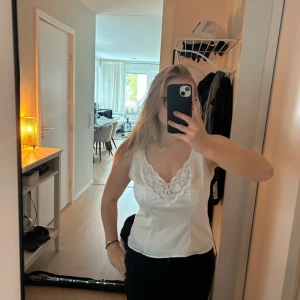 Vit spetsdetaljerad linnetopp - Säljer en vit linnetopp med vacker spets runt v-ringad halsringning och en liten rosett framtill. Toppen är ärmlös och har en mjuk, feminin siluett med lätt glansigt material. Perfekt för dig som gillar romantiska detaljer och en clean look.