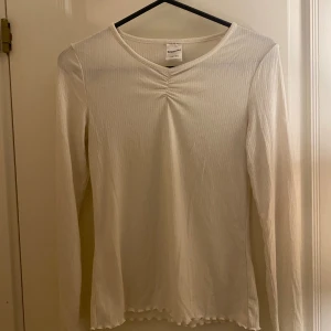 Vit långärmad topp Kappahl - Säljer en vit långärmad topp från Kappahl i storlek 158/164. Toppen har ribbad struktur, v-ringning med snygg rynkning framtill och vågig kant längst ner. Perfekt för dig som gillar enkel och stilren design.