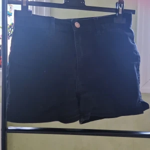 Svarta högmidjade shorts från H&M - Svarta shorts från H&M i storlek 152, med klassisk femficksdesign och knappstängning fram. Modellen är högmidjad och har en enkel, stilren look som passar perfekt till sommaren. Materialet är mjuk bomull med lite stretch för skön passform.