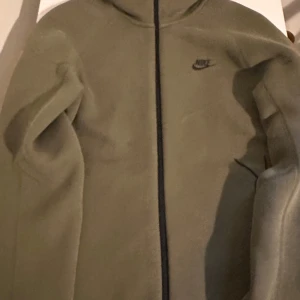 Olivgrön zip hoodie från Nike  - Olivgrön hoodiejacka från Nike med hel dragkedja framtill och svart Nike-logga på bröstet. Jackan har huva och är tillverkad i mjukt bomullsmaterial. Perfekt för chill dagar eller när du vill ha en sportig look.