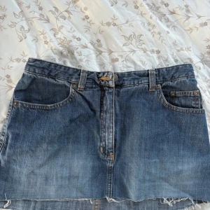 Jeanskjol från L.O.G.G. i blå denim - Snygg blå jeanskjol från L.O.G.G. i klassisk femficksmodell med bälteshällor och rå kant nertill. Kjolen har dragkedja och knapp framtill samt dekorativa bakfickor med broderade detaljer. Perfekt för dig som gillar en avslappnad och trendig stil.