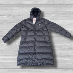 Lång Nike jacka - Nike dunjacka i svart med huva och Storm-FIT-teknologi. Jackan är lång, har två snedställda dragkedjefickor och är fodrad med Primaloft syntetfyllning. Perfekt för kalla dagar och har en clean look med diskret Nike-logga på bröstet. Lapparna är av, men följer med