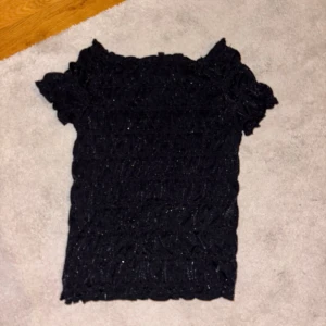 Svart glittrig topp med volangärm - Säljer en svart glittrig topp med korta volangärmar och smockad struktur. Toppen har en figurnära passform och är perfekt för dig som vill sticka ut lite extra. Materialet är stretchigt och bekvämt.