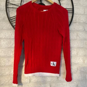 Röd stickad tröja från Calvin Klein - Riktigt snygg röd stickad tröja från Calvin Klein Jeans med vit ribbad kant vid ärmslut och nederkant. Klassisk kabelstickad design och stor logga framtill. Perfekt för dig som vill ha en färgstark och trendig look.