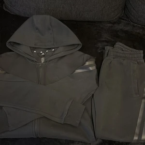 Svart hoodie och byxor ( SET) - Svart hoodie från Adidas med dragkedja och huva, klassiska svarta ränder på ärmarna och Adidas-logga på bröstet. Tröjan är långärmad och har ribbade muddar. Materialet är mjuk bomull och polyester, perfekt för chill eller träning.