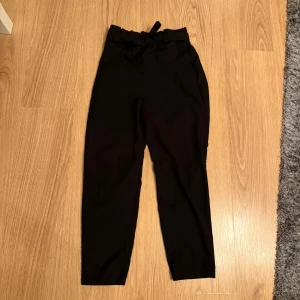 Svarta högmidjade byxor med knyt - Snygga svarta byxor med hög midja och ett trendigt knytband i midjan. Byxorna har en rak passform och är tillverkade i ett mjukt tyg som känns skönt mot huden. Perfekta för dig som gillar stilrena och moderna plagg med en twist.