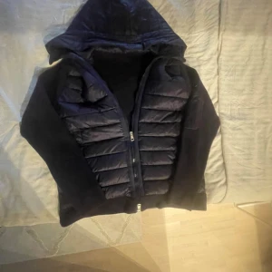 Mörkblå Moncler pufferjacka med huva - Snygg mörkblå pufferjacka från Moncler med stickade ärmar och huva. Jackan har quiltad front, dragkedja och Moncler-logga på ärmen. Perfekt för kyliga dagar och ger en riktigt clean look.