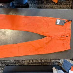 Orange jeans från Dondup, storlek 30 - Snygga orange jeans från Dondup i storlek 30. Modellen har klassisk femficksdesign, smal passform och coola slitningar på benen. Jeansen är tillverkade i bomull och har knappar i midjan. Perfekt för dig som vill sticka ut med färg.
