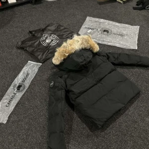 Svart dunjacka från Canada Goose - Säljer en svart dunjacka från Canada Goose med avtagbar huva och snygg pälskant. Jackan har klassisk patch på ärmen, dragkedja och knappar framtill samt flera fickor. Perfekt för kalla dagar och riktigt stilren look.