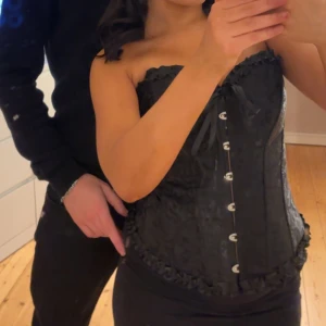 Svart korsett med snörning och knappar - Säljer en svart korsett med snygga knappar framtill och dekorativ snörning upptill. Korsetten har en lätt glansig yta och fina volangdetaljer längs kanterna. Den är i storlek M, men den går att göra mindre med snörerna!