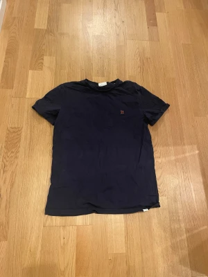 Mörkblå t-shirt från Les Deux - Snygg mörkblå t-shirt från Les Deux i storlek M. Klassisk passform med korta ärmar och rund hals. Diskret röd broderad detalj med 'II' på bröstet. Tillverkad i mjuk bomull som känns skön mot huden.