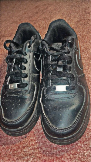 Svarta Nike Air Force 1 sneakers - Säljer ett par klassiska svarta Nike Air Force 1 sneakers i skinn med låg profil och rund tå. Skorna har snörning och en platt sula med det ikoniska Nike Swoosh på sidan. Perfekt för dig som gillar streetstyle och vill ha en tidlös look.