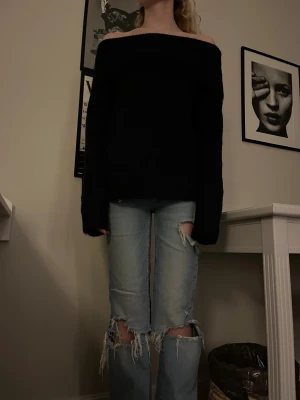 Svart offshoulder stickad tröja - Säljer en svart stickad tröja med offshoulder-design och bred ribbad kant upptill. Tröjan har lång ärm och en loose passform som ger en chill vibe. Perfekt att styla med slitna jeans eller kjol för en cool look.