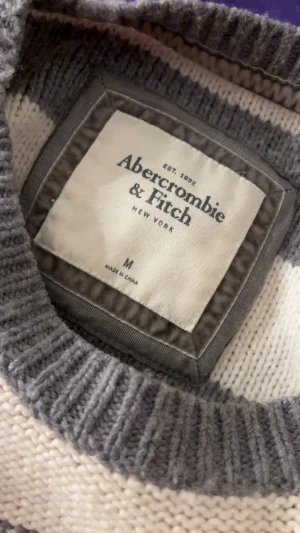 Randig stickad Abercrombie & Fitch - Snygg stickad tröja från Abercrombie & Fitch i storlek M. Tröjan har breda horisontella ränder i grått och beige, rund hals och ribbade muddar vid ärmslut och nederkant. Perfekt för dig som gillar klassisk och chill stil. (Nopprig)