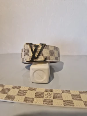 Louis Vuitton rutig beige bälte - Säljer ett snyggt bälte från Louis Vuitton med klassiskt rutigt mönster i beige och brunt. Bältet har en ikonisk LV-spänne i metall och är tillverkat i skinn. Perfekt för att lyfta din outfit med lite lyxig vibe.