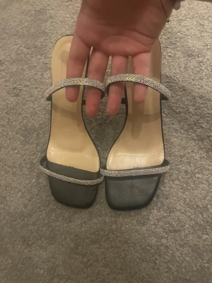 Svarta klackar med glittriga remmar - Säljer ett par svarta klackar från Shein i storlek 39. De har två smala remmar med glittriga paljettdetaljer och en fyrkantig tå. Klacken är smal och ger en elegant look. Perfekt för dig som gillar stilrena och trendiga skor med lite extra bling.