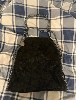 Svart handväska med kedjedetaljer - Snygg svart handväska i mjuk sammet med silverfärgade kedjor som axelremmar och runt kanterna. Väskan har en dragkedja på baksidan och rymligt innerfack. Perfekt för dig som gillar en cool och unik stil.