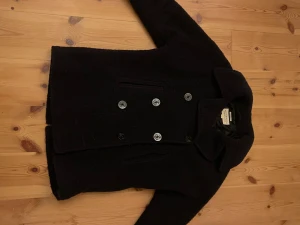 Mörkblå skepparkavaj från Ralph Lauren - Klassisk mörkblå skepparkavaj från Ralph Lauren. Jackan har knäppning med snygga metallknappar med ankare, bred krage och är tillverkad i ull. Perfekt för dig som vill ha en tidlös och stilren look. Lite nopprig men annars mycket bra skick