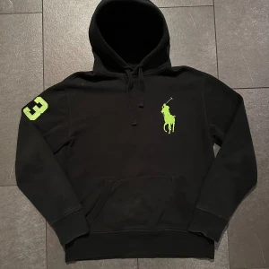 Svart hoodie Polo Ralph Lauren M - Svart hoodie från Polo Ralph Lauren med stor neongrön polospelare broderad på bröstet och en neongrön siffra 3 på ärmen. Klassisk känguruficka, snörning i huvan och mjukt material i bomull och polyester. Perfekt för en chill och sportig look.