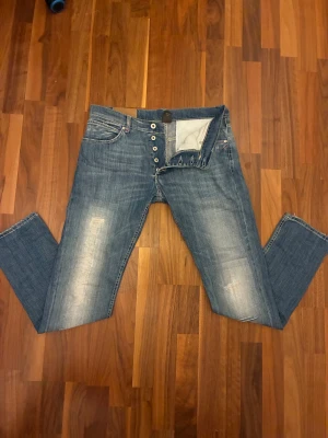 Dondup - Säljer ett par Dondup jeans i ljusblå tvätt med snygga slitningar och knappar framtill. Jeansen har tapered passform, klassiska fem fickor och cool detalj med broderad bakficka. Perfekt för dig som gillar street och avslappnad stil