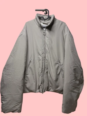Grå pufferjacka från Acne Studios storlek 50 (L) - Säljer en oversized grå pufferjacka från Acne Studios med hög krage och rund metallring på dragkedjan. Jackan har två snedställda fickor med dragkedja och är tillverkad i syntetmaterial. Perfekt för dig som gillar minimalistisk och clean stil.